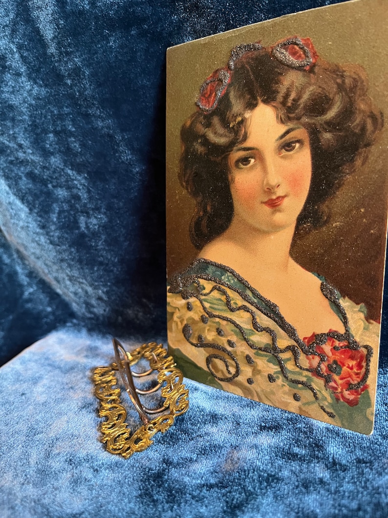 Op de afbeelding: Een gouden haarspeld met een decoratief ontwerp. De speld ligt op een blauw fluwelen oppervlak. Op de achtergrond is een vintage ansichtkaart te zien van een vrouw met donker haar en een blauw-witte jurk.