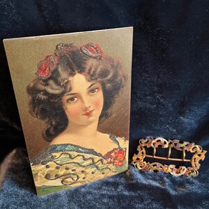Op de afbeelding: Vintage ansichtkaart met een vrouw met donker haar en rode bloemen in haar haar. Ze draagt een gepatroneerd jurkje met een zwarte kantversiering. De ansichtkaart heeft een gouden achtergrond.