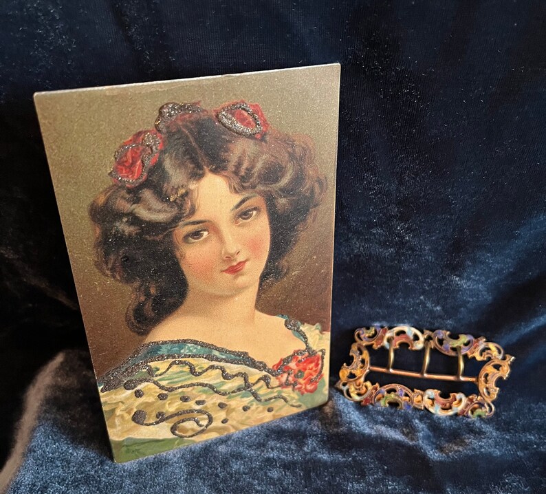 Op de afbeelding: Vintage ansichtkaart met een vrouw met donker haar en rode bloemen in haar haar. Ze draagt een gepatroneerd jurkje met een zwarte kantversiering. De ansichtkaart heeft een gouden achtergrond.