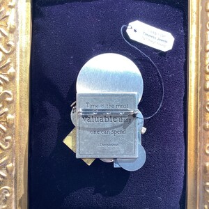 May include: A silver-toned metal pin with a quote about time being the most valuable thing one can spend. The quote is attributed to Theophrastus. The pin is framed in a gold-colored frame.