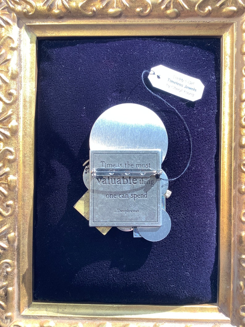 May include: A silver-toned metal pin with a quote about time being the most valuable thing one can spend. The quote is attributed to Theophrastus. The pin is framed in a gold-colored frame.