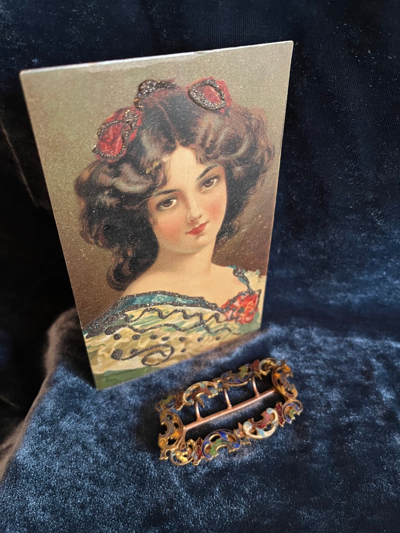 Op de afbeelding: Een vintage, sierlijke, goudkleurige metalen gesp met blauwe, rode en groene emaille accenten. De gesp ligt op een donkerblauw fluwelen oppervlak.