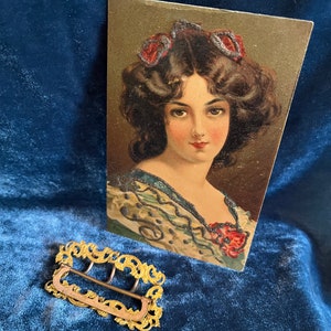 Op de afbeelding: Een gouden metalen gesp met een sierlijk ontwerp. De gesp ligt op een blauw fluwelen oppervlak. Op de achtergrond is een vintage ansichtkaart te zien met een vrouw met donker haar en een bloemenjurk.