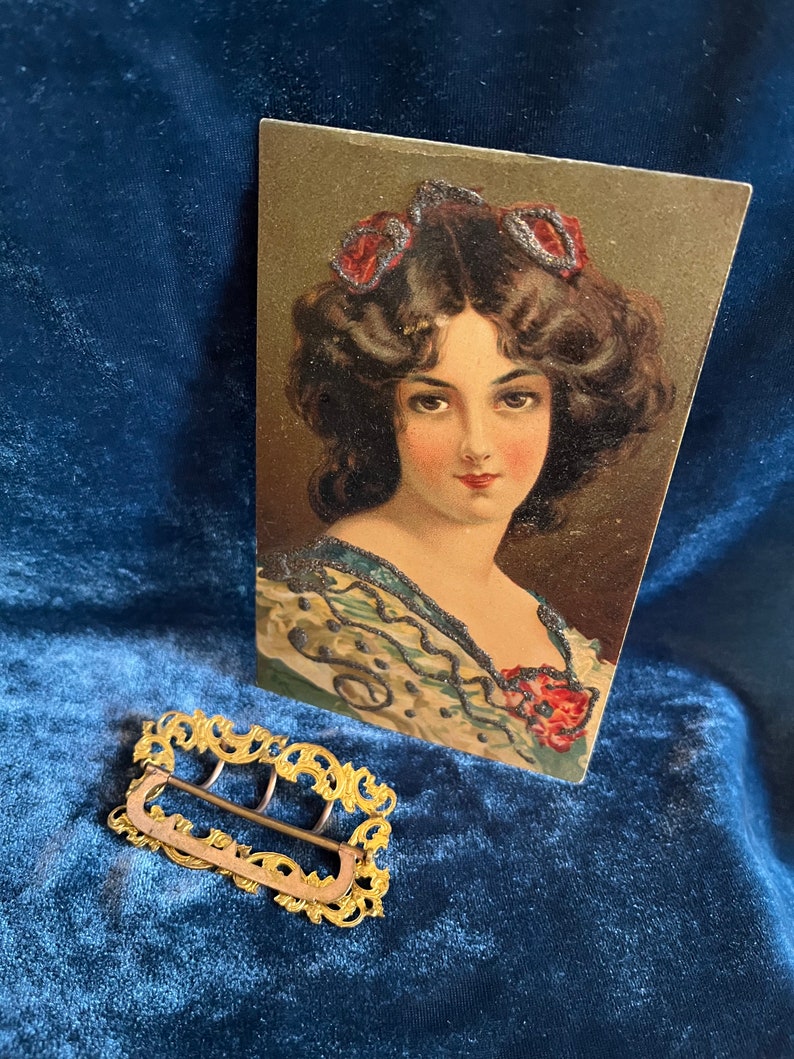 Op de afbeelding: Een gouden metalen gesp met een sierlijk ontwerp. De gesp ligt op een blauw fluwelen oppervlak. Op de achtergrond is een vintage ansichtkaart te zien met een vrouw met donker haar en een bloemenjurk.