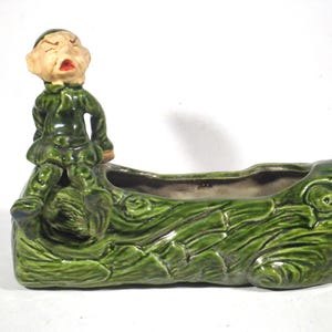May include: A vintage ceramic planter featuring a green log design with a seated figure. The figure has a tan face, red mouth, and is dressed in a green outfit. The planter is a rectangular shape.