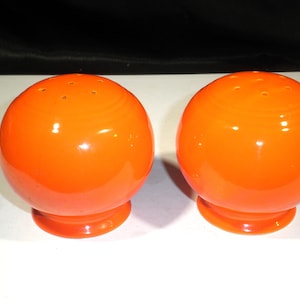 May include: Pair of bright orange, spherical salt and pepper shakers. Each shaker has a round base and a series of small holes on top. The shakers are made of a glossy material and are set against a white surface.