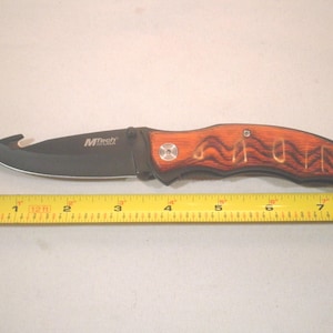 May include: A brown and black folding pocket knife with a wood handle. The handle has the word "MOM" carved into it. The knife is 7 inches long.
