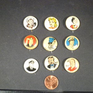 May include: A collection of vintage pinback buttons featuring cartoon characters and nautical themes. The buttons are round, with colorful illustrations and metal pin backs. A penny is included for size reference.