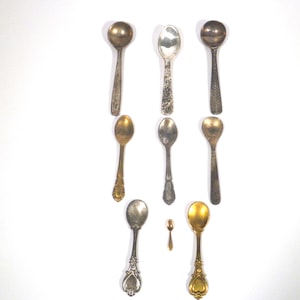 May include: A collection of antique spoons in silver and gold tones. The spoons have different handle designs, some ornate, some plain. The spoons are arranged on a white background. Measurements are in centimetres.