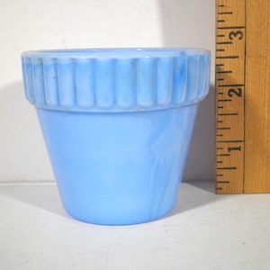 May include: A light blue, small ceramic flower pot with a ribbed rim. The pot is approximately 2.5 inches tall and has a tapered shape. The pot is next to a wooden ruler.