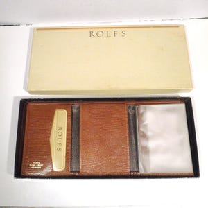 May include: A brown leather wallet with a textured finish, presented in an off-white box. The wallet is open, revealing card slots and a clear ID window. The box lid displays the brand name "ROLFS".