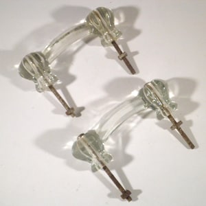 May include: Two clear glass drawer pulls with a curved, arched design. Each handle features ribbed detailing at the ends and includes two attached metal bolts with nuts extending from the base for mounting purposes on furniture or cabinetry.