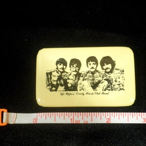 Peut inclure: Une épingle rectangulaire jaune avec une image en noir et blanc des Beatles, avec le texte "Sgt. Pepper's Lonely Hearts Club Band".