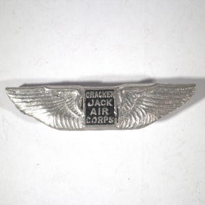 May include: A silver-colored metal pin in the shape of wings, with the text "CRACKER JACK AIR CORPS" in a black rectangle at the center. The wings have detailed feathering.