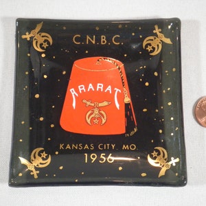 Op de afbeelding: Een zwarte glazen asbak met gouden accenten en een rode fez met de tekst "Ararat" en "C.N.B.C. Kansas City, Mo. 1956".