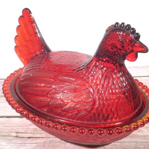 May include: A translucent red glass hen on a nest dish. The lid is shaped like a sitting hen with detailed feather textures, while the base features a beaded rim design. The entire piece is crafted from vibrant, deep red pressed glass.