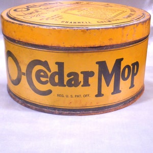 May include: Vintage O-Cedar Mop tin with a yellow and black color scheme. The cylindrical container features the words "O-Cedar Mop" in large black letters. The top of the tin has additional text and a label. The container is slightly rusted.