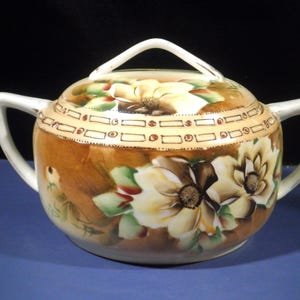 May include: A round, lidded sugar bowl with two side handles and a top handle. The bowl is painted with brown, cream, and green floral designs. The rim of the bowl features a decorative band.