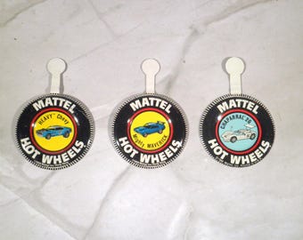 May include: Three circular metal pin-back badges featuring the Mattel Hot Wheels logo in black and white. Each badge displays a vintage car illustration: a blue Heavy Chevy, a blue Mighty Maverick, and a white Chaparral 2G, each with a small tab.