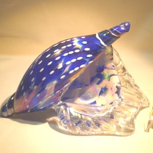 May include: A blue glass seashell with white polka dots and iridescent accents sits on a clear glass base.