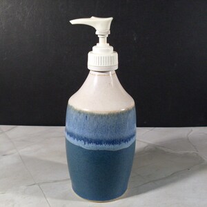 May include: A ceramic soap dispenser with a white plastic pump mechanism. The bottle features a tapered neck and a cylindrical body with a two-tone glaze, transitioning from a creamy off-white at the top to a deep, speckled blue at the base.