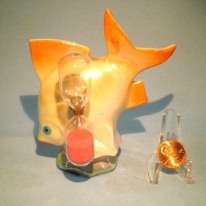 May include: A ceramic fish figurine with a sand timer inside. The fish is painted in shades of yellow, orange, and white. The sand timer is filled with red sand.