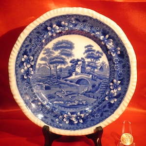 May include: A blue and white porcelain plate with a floral border and a scene of a bridge over a stream with trees and a small house in the background.
