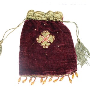 May include: A deep burgundy velvet drawstring pouch featuring a gold metallic top trim and matching gold cord drawstrings with tassels. The front is decorated with a central gold embroidered cross-shaped motif and a bottom fringe of amber beads.