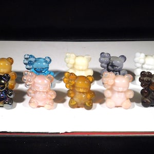 Puede incluir: Una colección de pequeñas figuras de osos en varios colores, incluyendo dorado, azul, rosa, amarillo, gris y marrón. Cada oso tiene un diseño único y se exhibe sobre una superficie blanca. Las figuras son probablemente objetos de colección o decorativos.