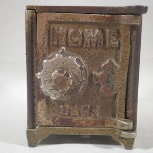 May include: A vintage cast iron home safe with a combination lock and a keyhole. The safe is painted in a rusty brown color and has the words "HOME SAFE" embossed on the front.