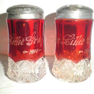 May include: A pair of vintage red glass salt and pepper shakers. Each shaker has a silver metal lid and a clear, cut-glass base. The red glass is etched with the name "Lillie Gray" and the year "1905".