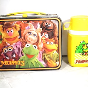 May include: Vintage metal lunchbox featuring The Muppets characters, with a yellow handle and trim. A matching yellow thermos with a Kermit the Frog graphic is also included. The lunchbox and thermos are a nostalgic collectible.