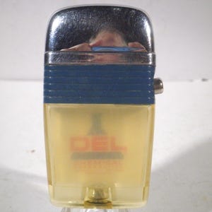 May include: A vintage lighter with a chrome top, a blue band, and a clear, yellow-tinted body. The lighter has the word "DEL" printed on the front in red and orange. A small metal button is on the side.