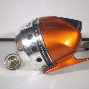 May include: A spincast fishing reel featuring a metallic silver front cone with circular ventilation holes and a glossy orange rear housing. The black band around the center displays the text CATCH MORE FISH. A coiled metal spring extends from the front.