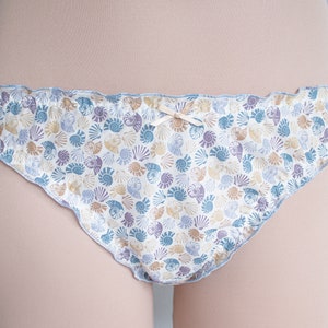 May include: White cotton underwear with a blue and brown seashell print. The underwear has a ruffled edge and a small bow at the centre.