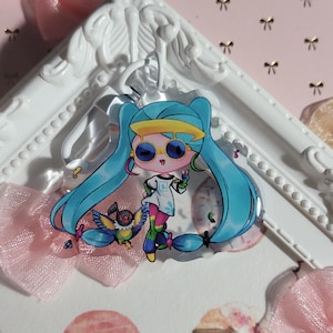 May include: A blue and yellow anime character with long blue hair and sunglasses. The character is holding a small bird in their hand. The character is on a clear acrylic keychain.