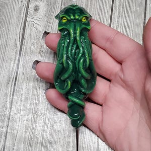 Peut inclure: Épingle verte sur le thème de Cthulhu avec des yeux jaunes et des tentacules. L'épingle est faite d'un matériau brillant et possède une longue et fine aiguille noire. Le design est détaillé et a un aspect texturé.