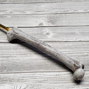 May include: A crochet hook with a bone-shaped handle. The handle is painted white and gray, with a textured appearance. The hook itself is gold-colored. The hook is lying on a wooden surface.
