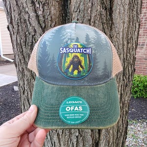 May include: A green and tan trucker hat featuring a forest scene with a Sasquatch graphic. The hat has a curved brim and a mesh back. The text "THE SASQUATCH!" is displayed on the front.