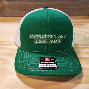 May include: Green and white trucker hat with the text "MAKE GREENLAND GREAT AGAIN". The hat features a green front panel and peak, with a white mesh back. The Richardson brand tag is visible.
