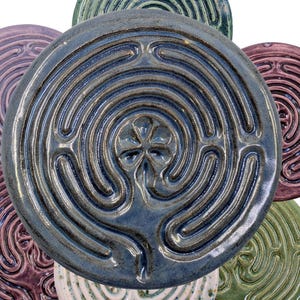 May include: A selection of ceramic labyrinth tiles in shades of blue, green, purple, and white. Each tile displays a concentric circular path design. The tiles are arranged in a flower-like pattern, highlighting their unique textures and glazes.