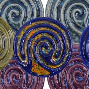 May include: A selection of ceramic plates featuring a spiral pattern. The plates are presented in a range of colours, such as blue, green, purple, and yellow. These round plates have a glossy finish and are arranged in a circular formation.