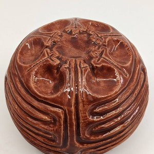 May include: A brown ceramic bowl with a unique, intricate design. The bowl has a rounded shape and a textured surface. The design features a series of raised lines and curves that create a complex pattern.