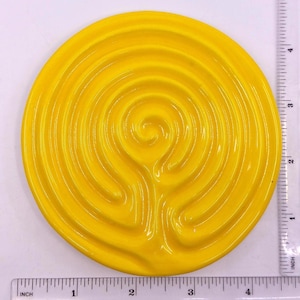 Op de afbeelding: Een heldergele keramische labyrint met een spiraalvormig ontwerp. Het cirkelvormige stuk heeft een diameter van ongeveer 10 cm. Het labyrint heeft een glanzende afwerking en staat tegen een witte achtergrond.