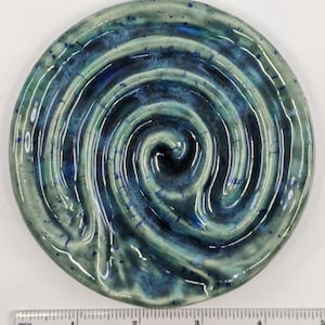 Puede incluir: Un posavasos redondo de cerámica con un esmalte azul y verde en espiral. El posavasos tiene un diseño texturizado en relieve que se asemeja a una espiral.