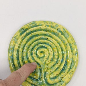 Op de afbeelding: Een ronde keramische onderzetter met een labyrintontwerp. De onderzetter heeft een spiraalpatroon in geel en blauwgroen, bespikkeld met donkere vlekken. Het ontwerp is gecentreerd.