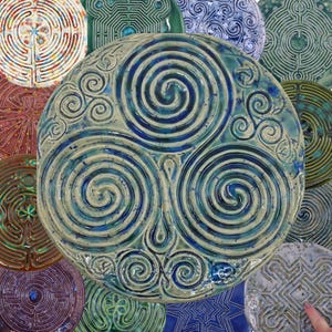 May include: A collection of ceramic plates with intricate designs. The central plate features a blue and green spiral pattern, surrounded by other plates with various labyrinth and geometric motifs. The plates have a glossy finish and are arranged in a circular composition.
