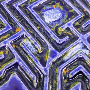 May include: Close-up of a blue ceramic tile with a raised geometric pattern. The pattern is outlined in black and white, with accents of yellow and orange.