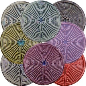 May include: A selection of ceramic labyrinth plates in shades of blue, green, yellow, purple, and red. Each plate displays a concentric circular path design with a central floral motif. The plates have a textured surface.