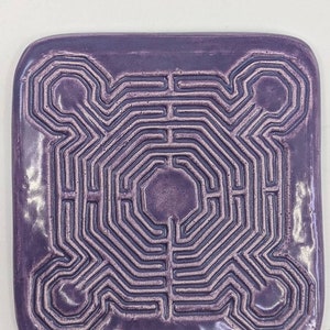 May include: A square ceramic tile with a purple glaze and a raised, embossed labyrinth design. The labyrinth is a complex pattern of lines and curves.
